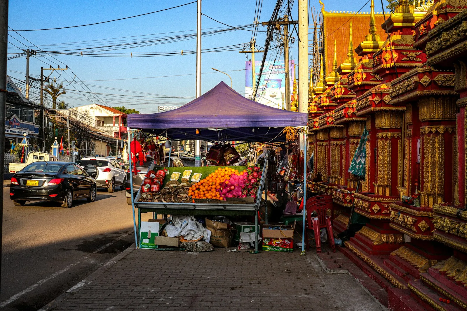 A fruit stall under a purple awning beside a gilded temple wall on a sunny street, with cars and power lines overhead.
