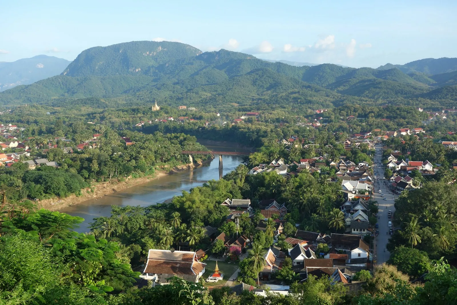 A wooded river bend and red-roofed houses below a green ridge, seen from a hilltop on a clear morning.