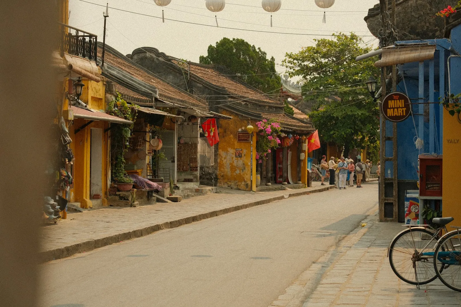 A quiet street lined with low yellow and blue shopfronts under hanging lanterns, with a bicycle parked at the kerb.