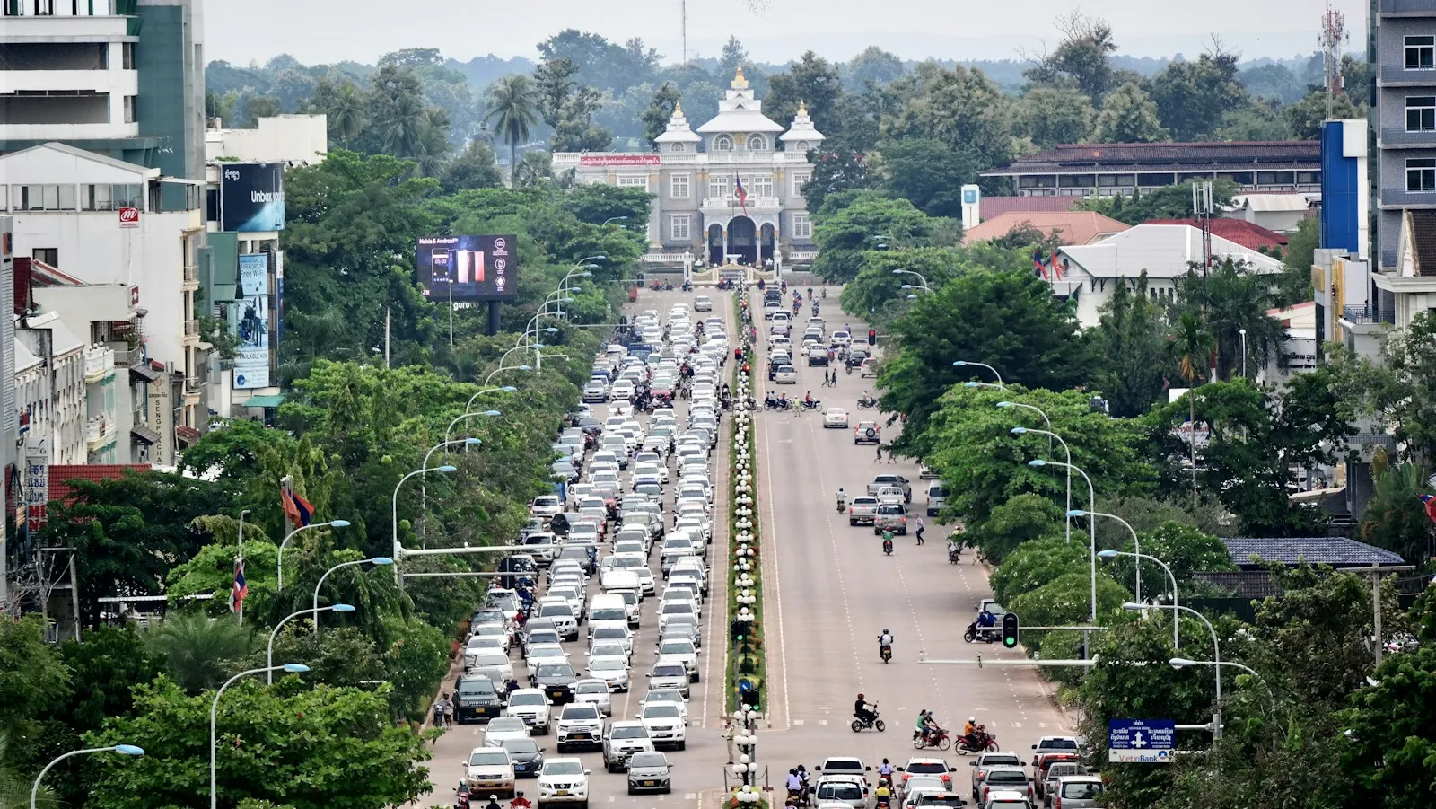 A wide avenue in Vientiane lined with cars and trees, leading to a pale gateway building at the far end.