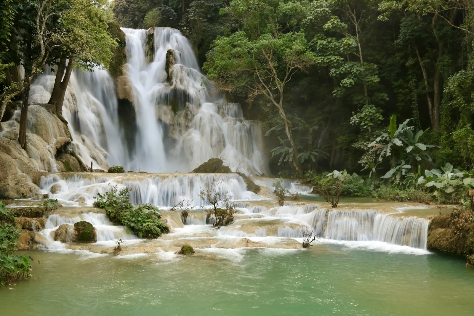 A tiered waterfall tumbling over pale rock into a turquoise pool, framed by green forest.