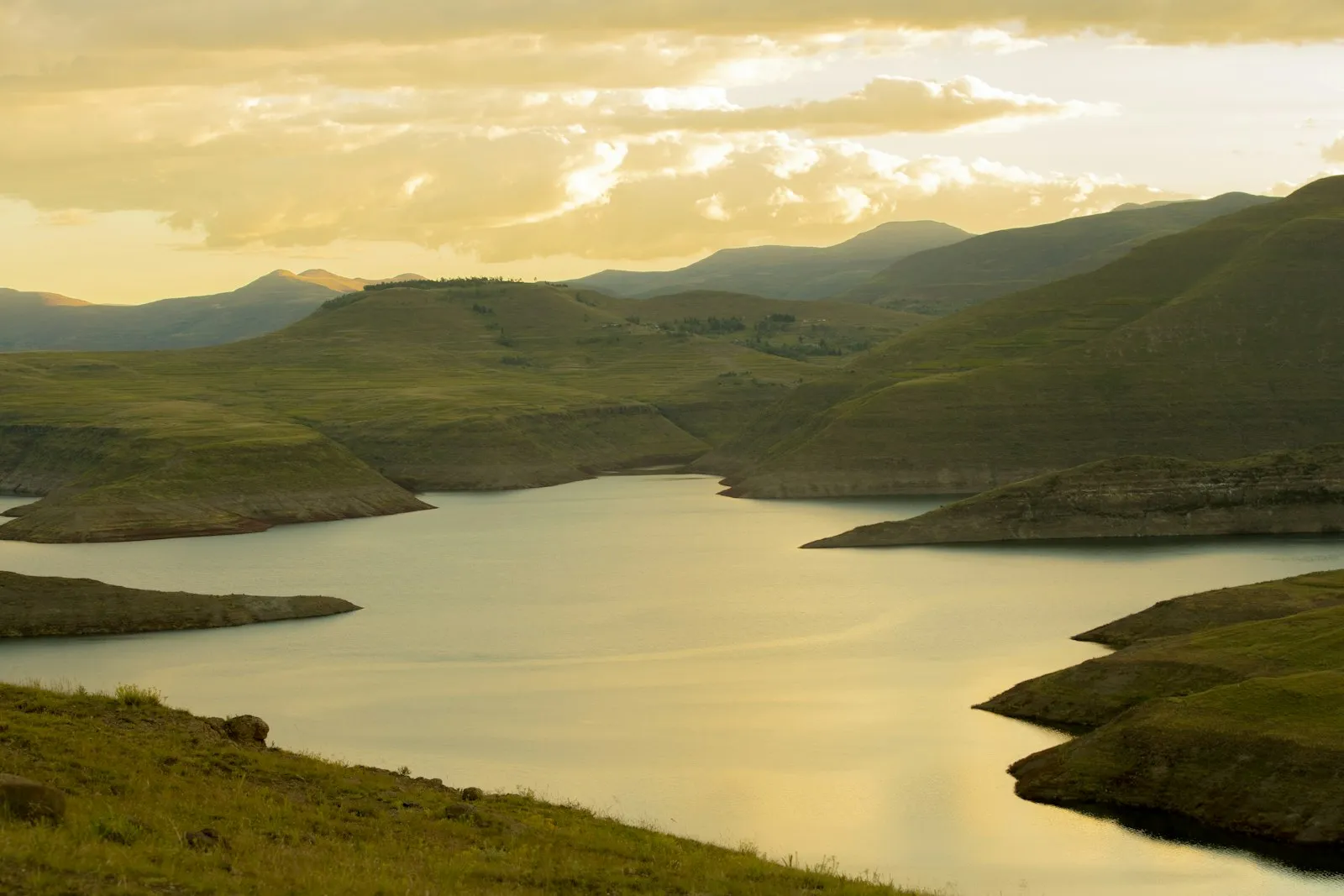Layered green hills beside a calm reservoir under a pale golden sky.
