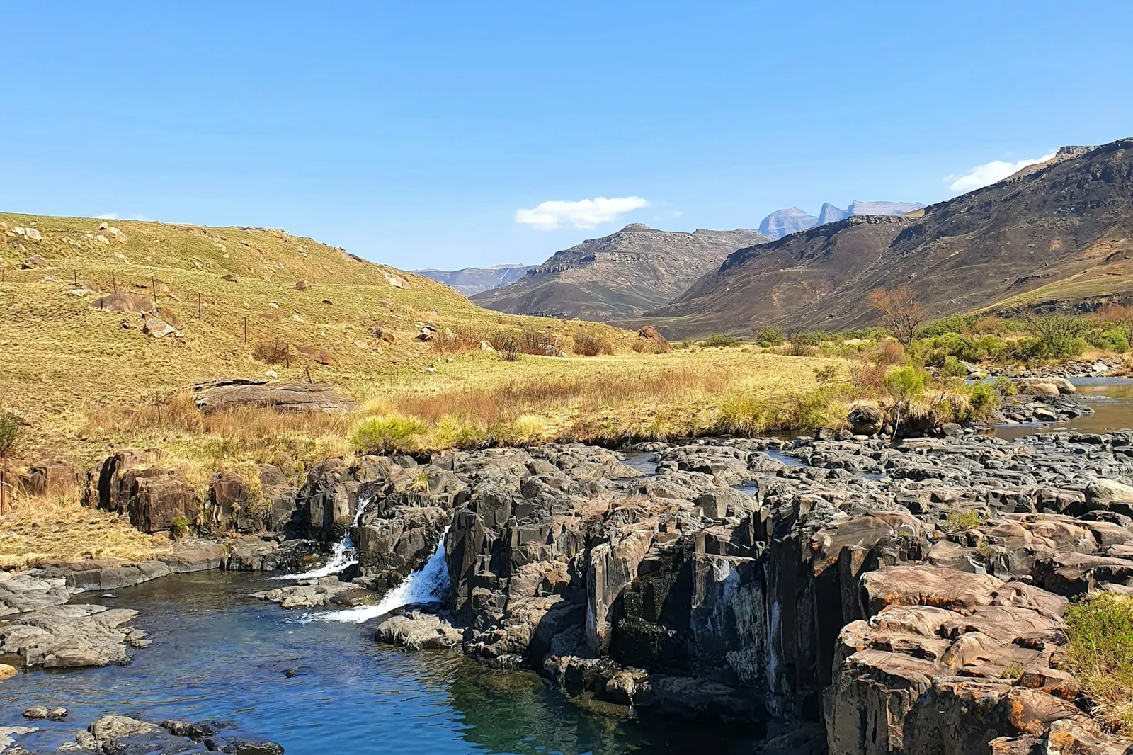 A small waterfall dropping into a blue pool between dark rock ledges, with a grassy valley and mountains behind.
