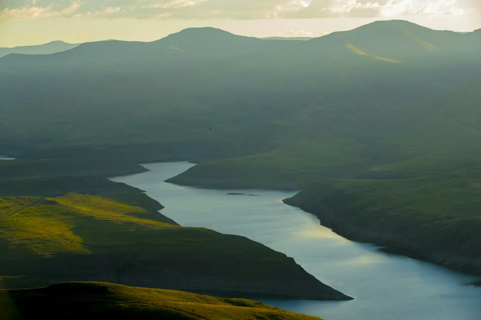 A blue-green reservoir winding between steep green mountain ridges in warm evening light.