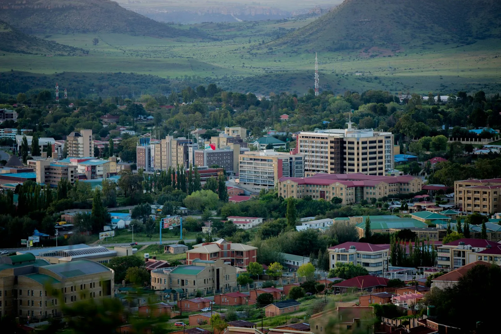 A hillside view over Maseru, with pale office blocks and red and green rooftops among trees, and green hills rising behind.
