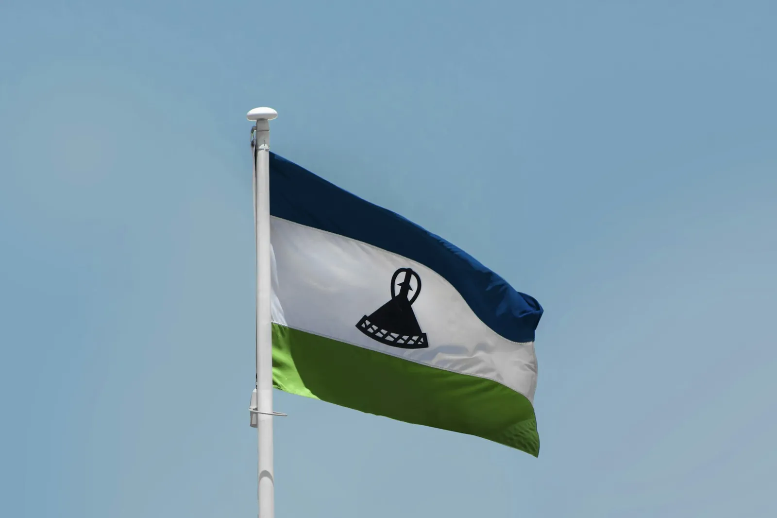 The blue, white and green flag of Lesotho flying from a white pole against a clear sky.