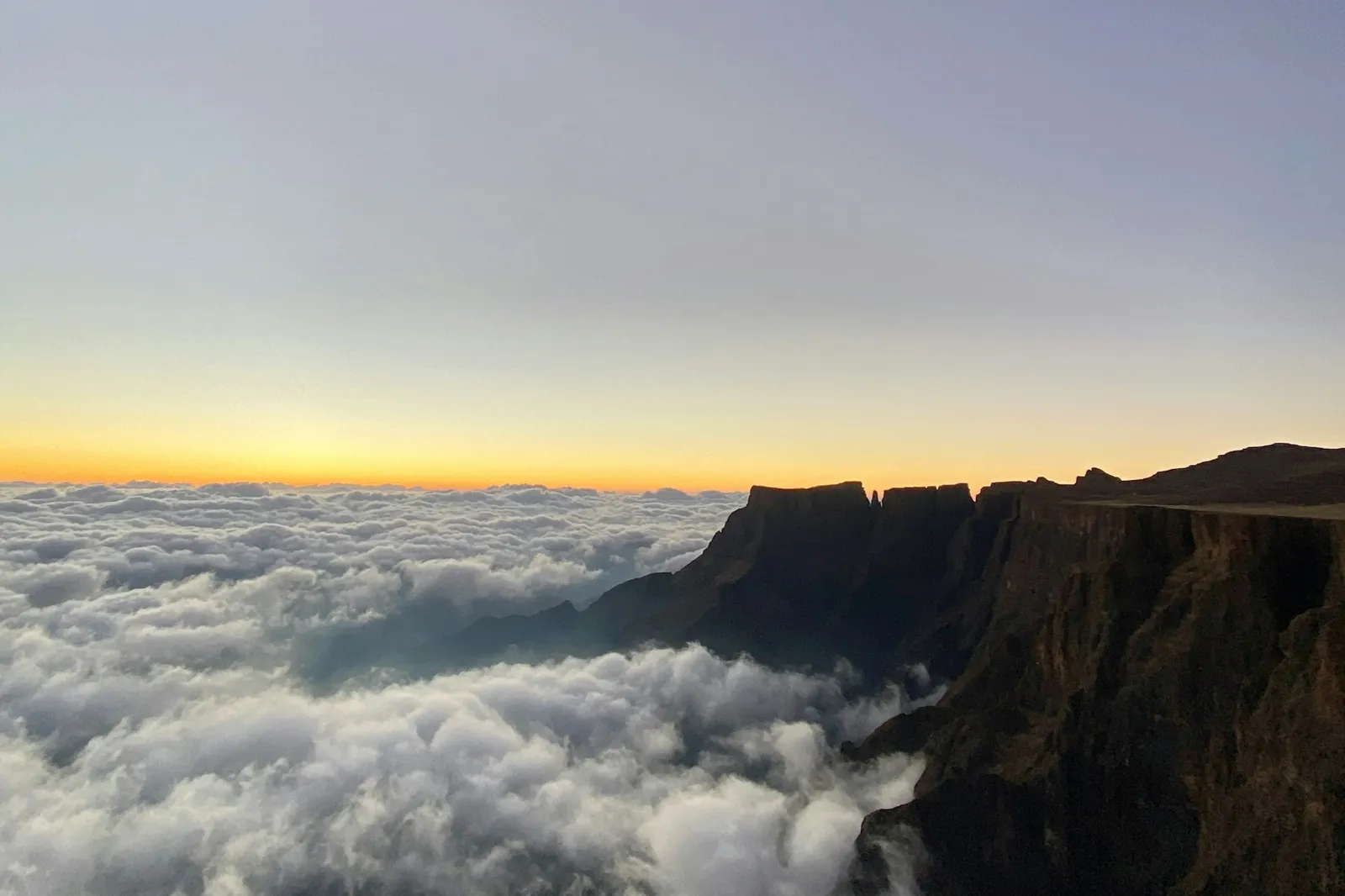 A sea of cloud filling a valley below a dark mountain ridge, with a band of orange light on the horizon.