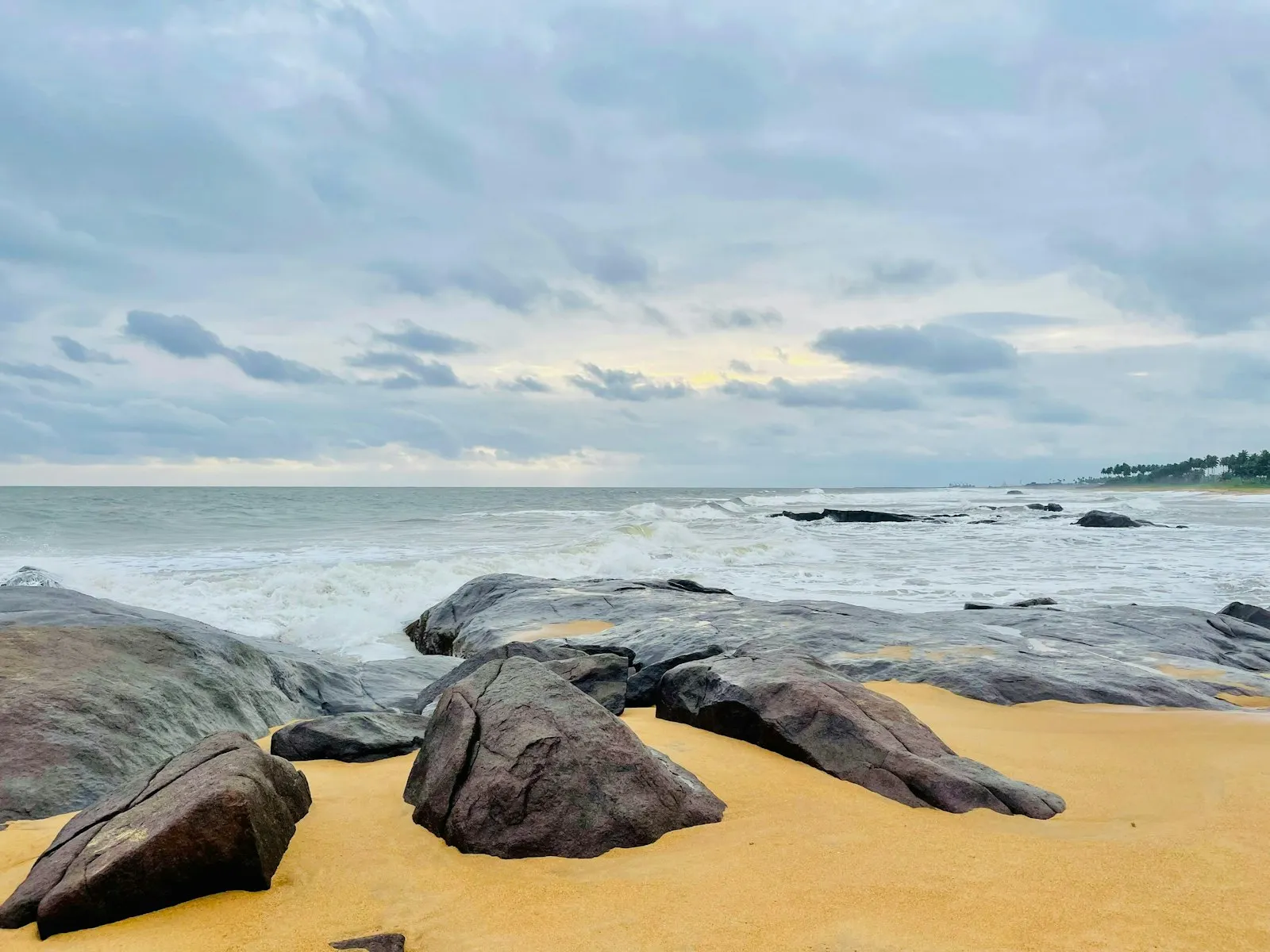 Dark rounded rocks on yellow sand with surf breaking behind them, under a grey and blue cloudy sky.