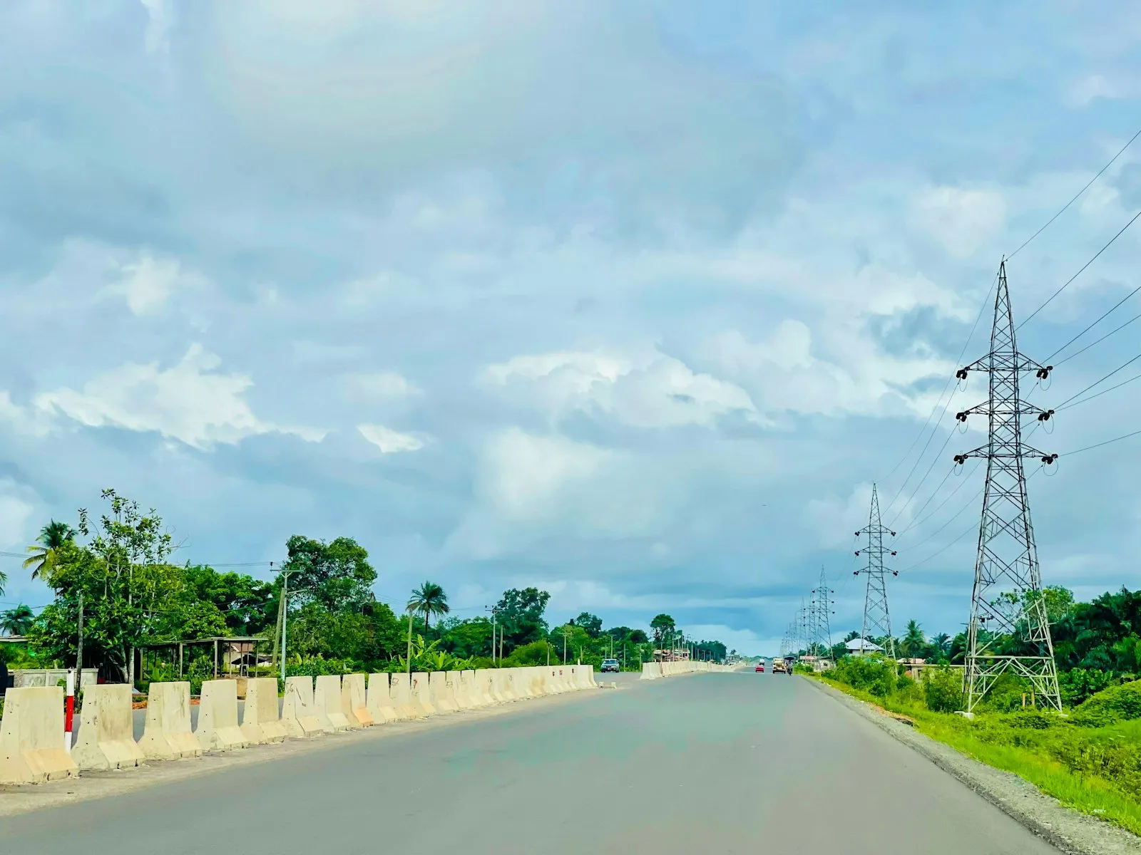 A wide paved road beside a low white wall and green trees, with an electricity pylon under a cloudy sky.