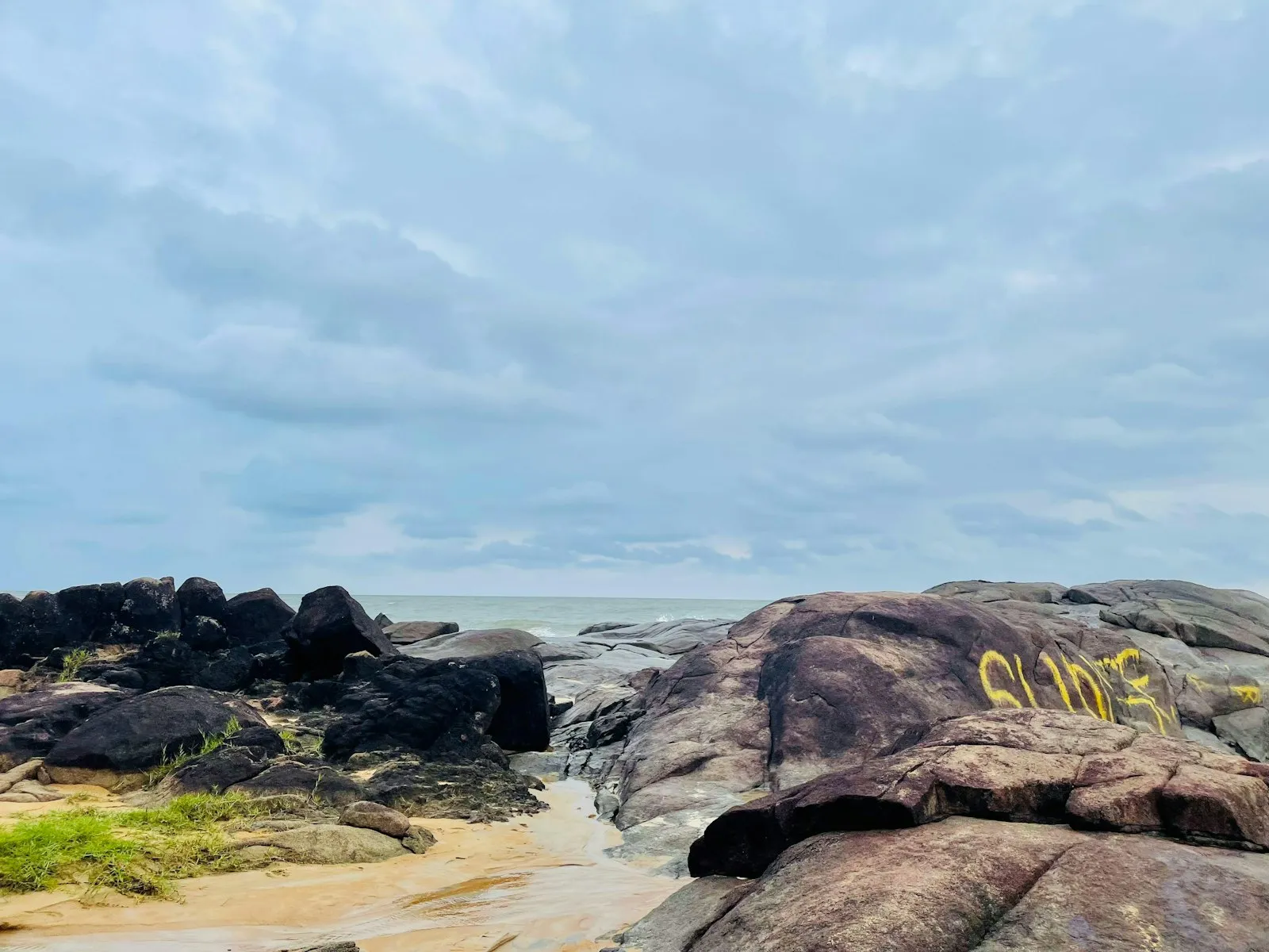 Weathered dark rocks and a patch of green grass on a sandy shore, under a pale blue cloudy sky.