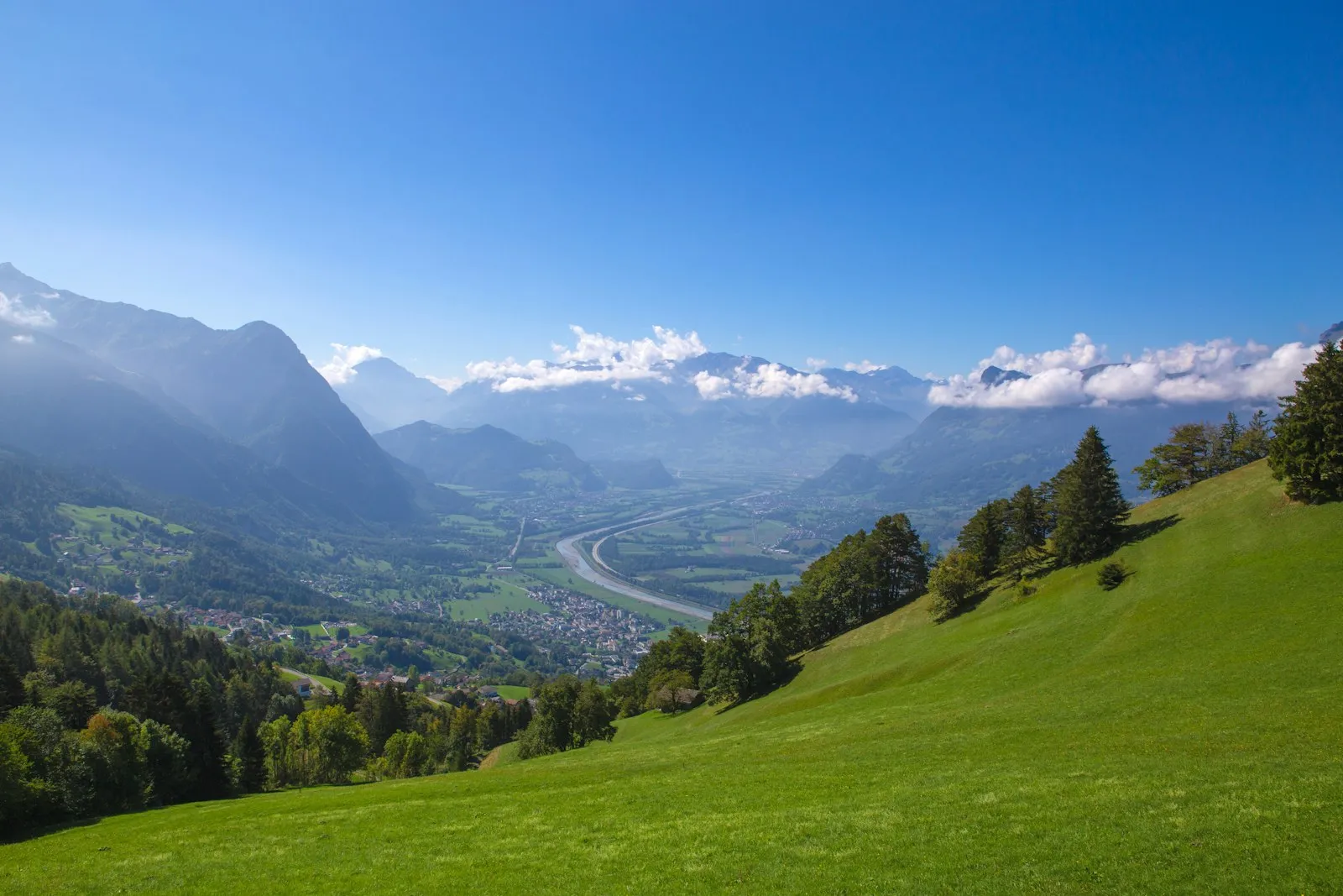 A green mountain slope above the Rhine valley and a small town in Liechtenstein, with hazy peaks beyond under a blue sky.