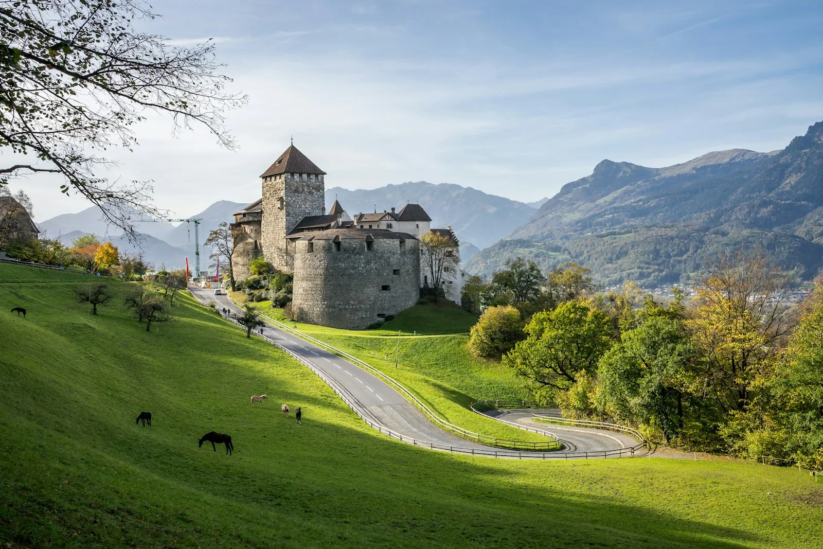 Vaduz Castle, a pale stone tower and walls on a green hillside above a winding road, with Alpine peaks behind it.
