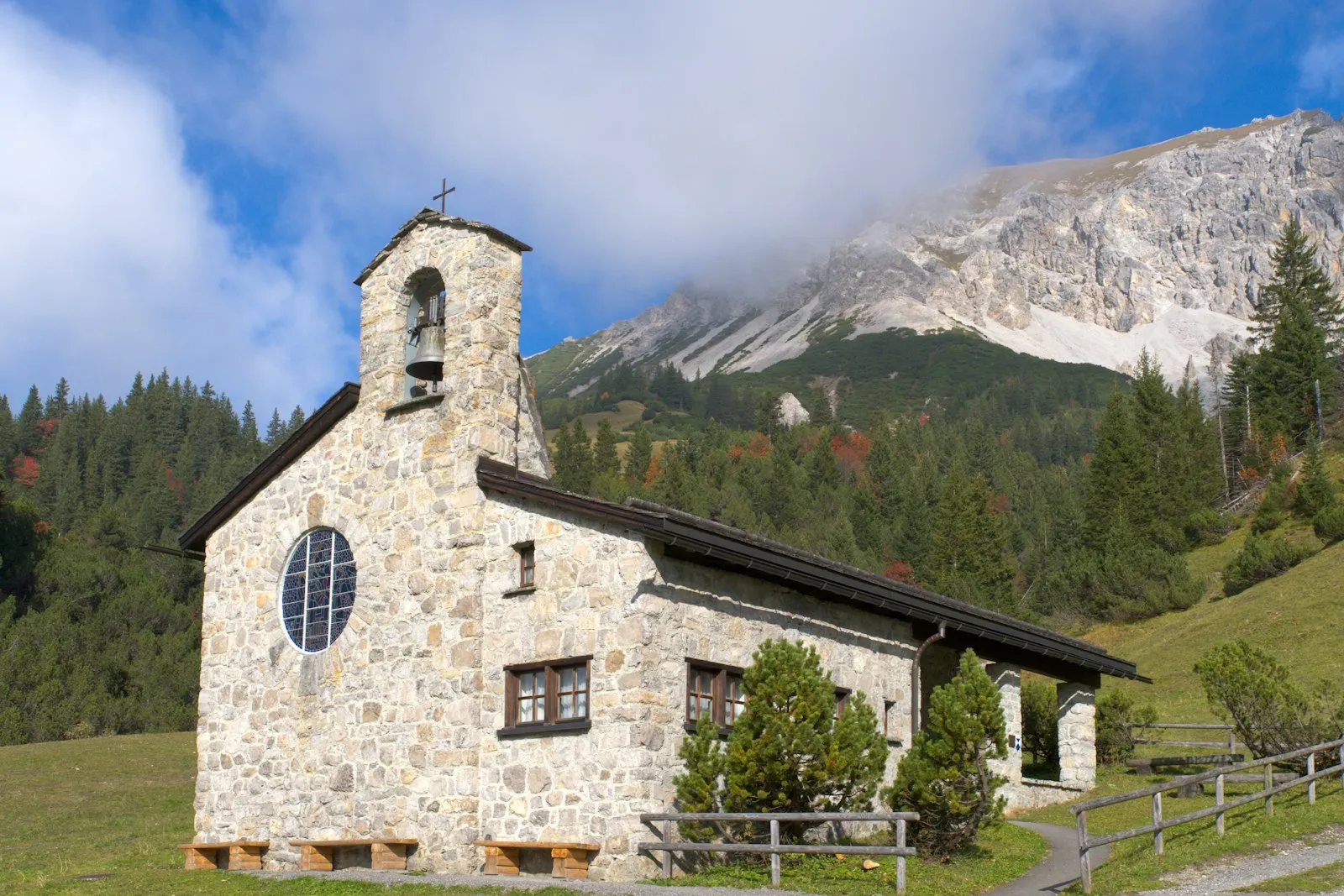 A small stone chapel with a bell gable and a round window below a cloudy mountain ridge in Malbun.