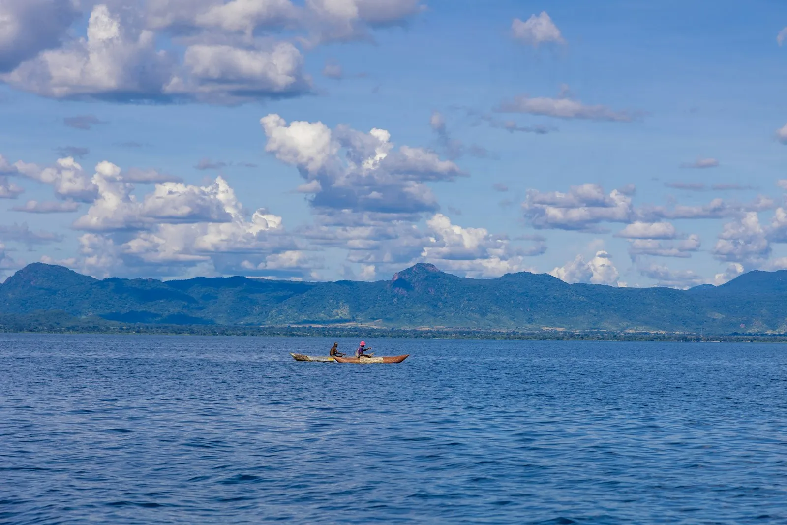 A small boat on the calm blue water of Lake Malawi, with a long blue mountain ridge across the far shore under scattered white clouds.