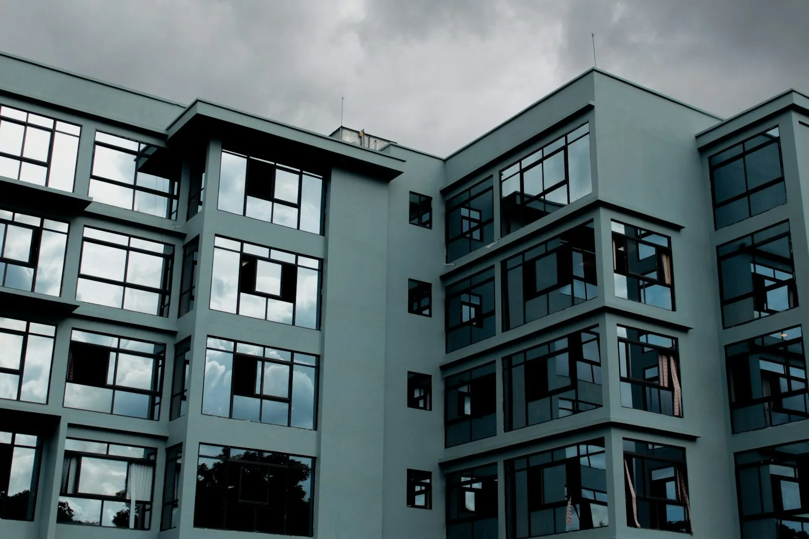 A modern five-storey apartment block with grey walls and large windows, seen from below against a cloudy sky.