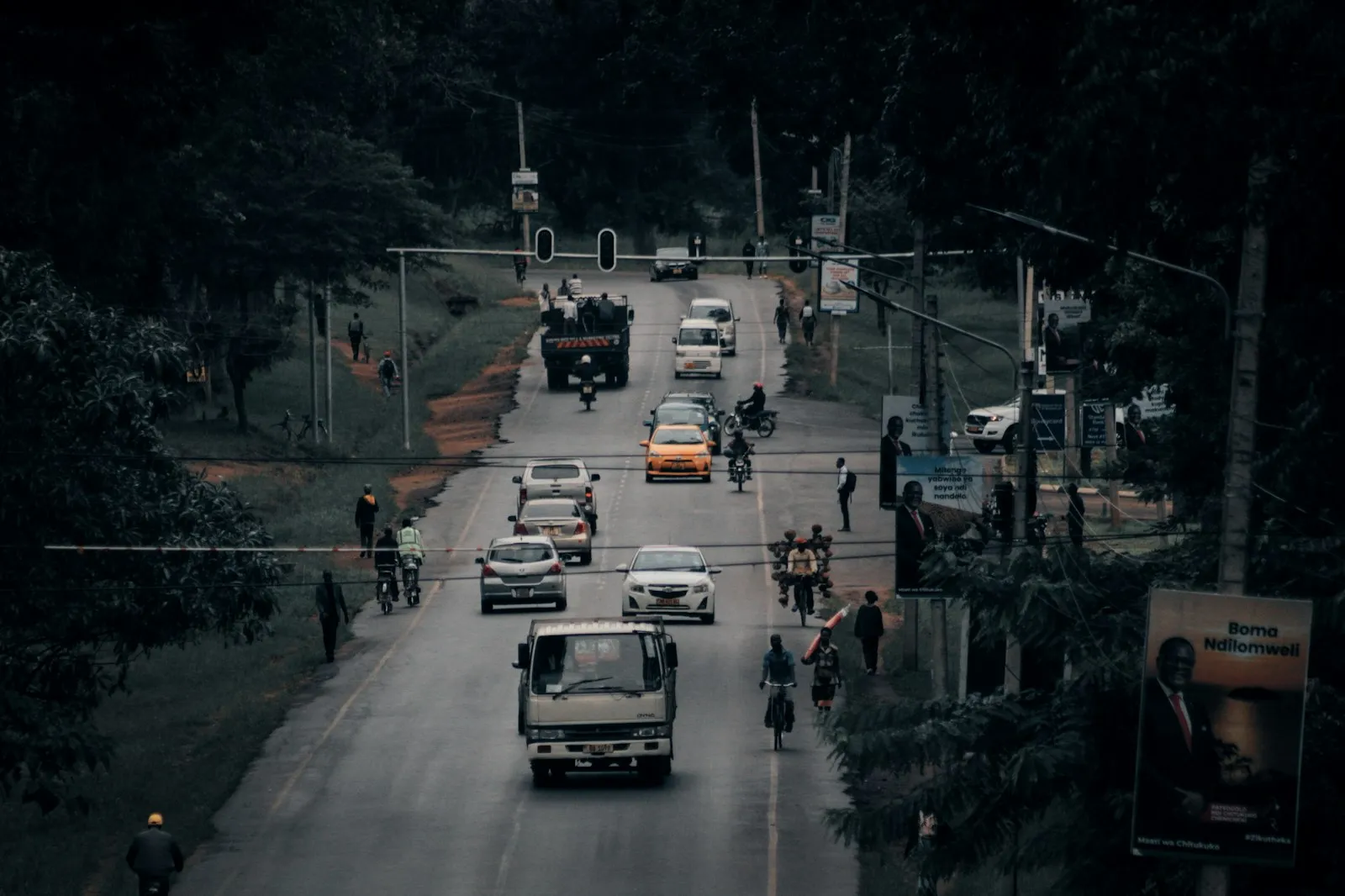 A busy tarred road in Lilongwe seen from above, with cars, a lorry and people walking beside it, and trees and billboards along the edge.