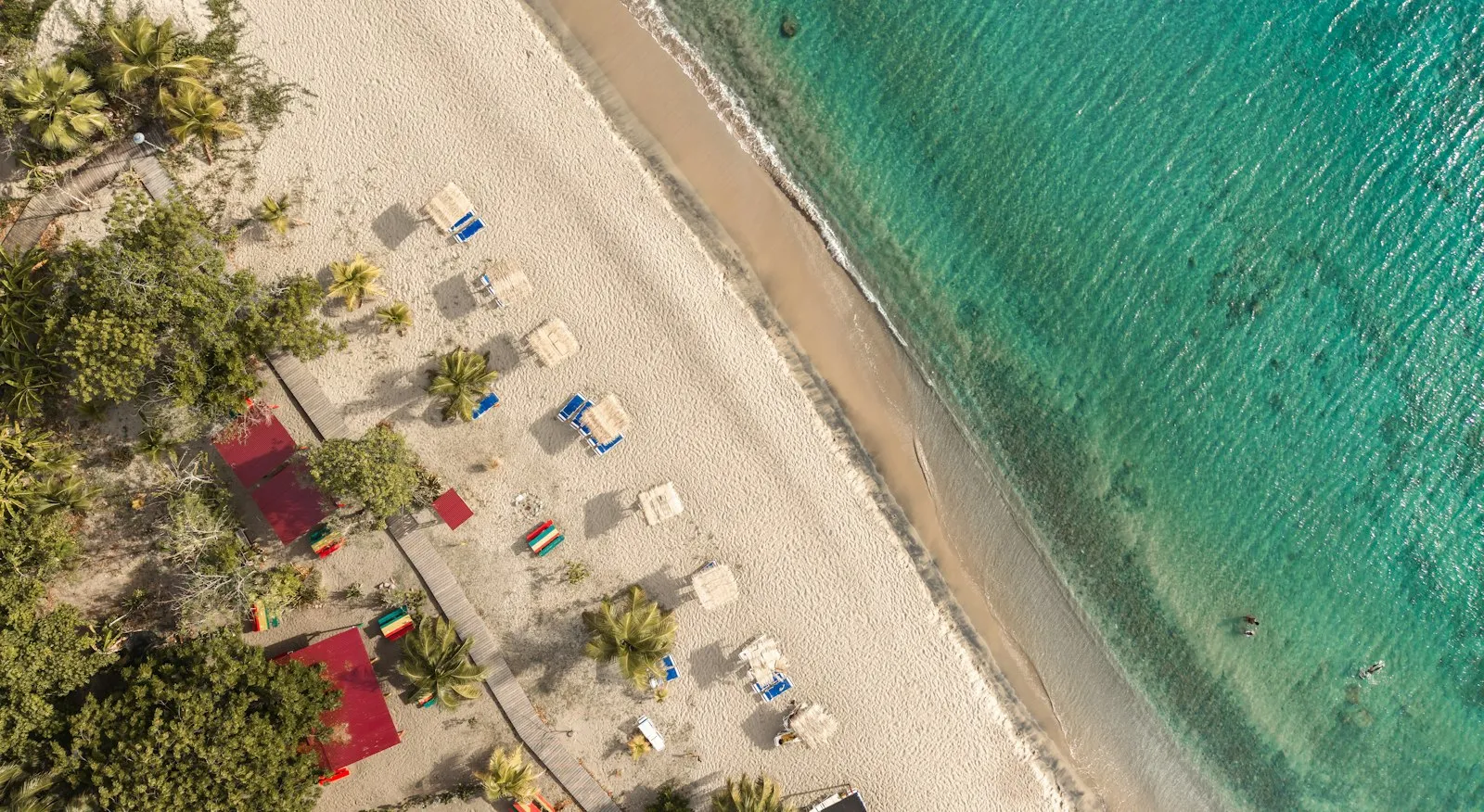 An aerial view of Pinney's Beach on Nevis, with loungers and palm trees along the pale sand beside turquoise water.