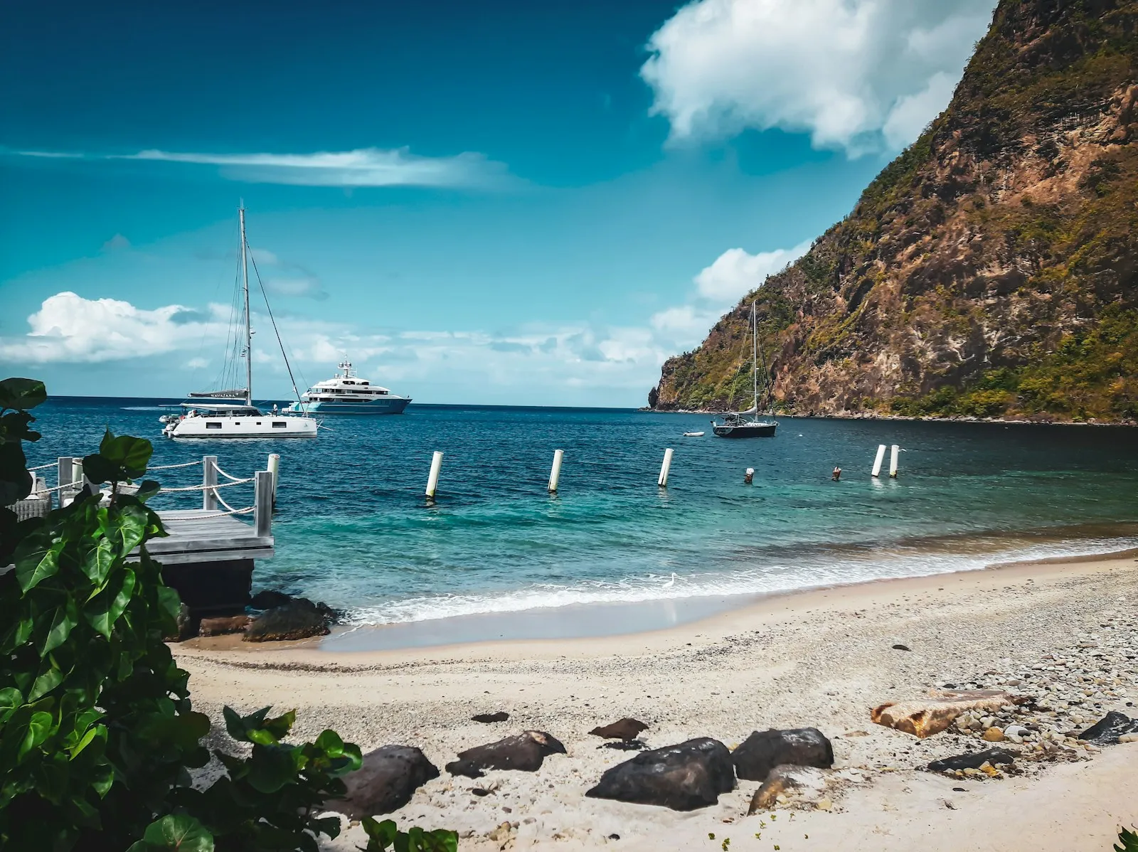 A beach with sailing boats at anchor and a steep green mountain behind, in Saint Lucia.