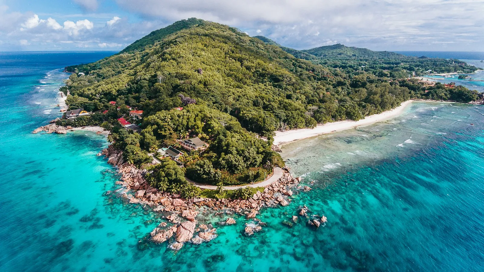 An aerial view of a green, forested island with a white beach edged by turquoise water, seen from above near La Digue, Seychelles.