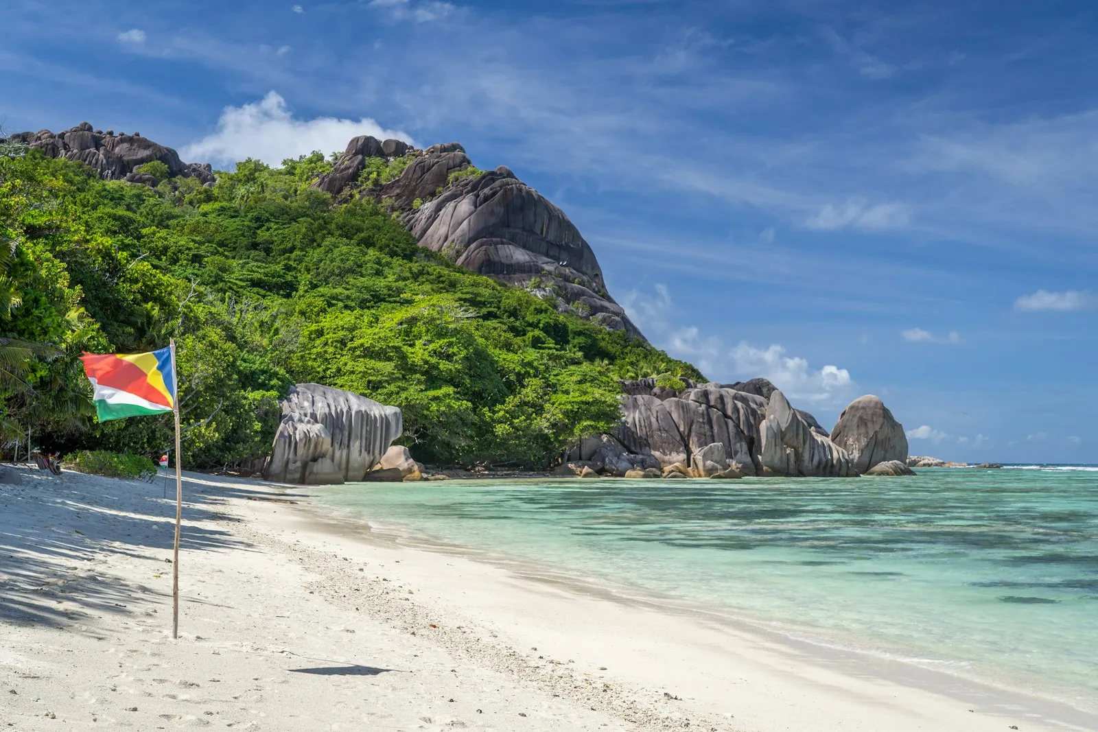 The Seychelles flag flying on a white sand beach beside granite boulders, green hills and turquoise water at Anse Source d'Argent on La Digue.