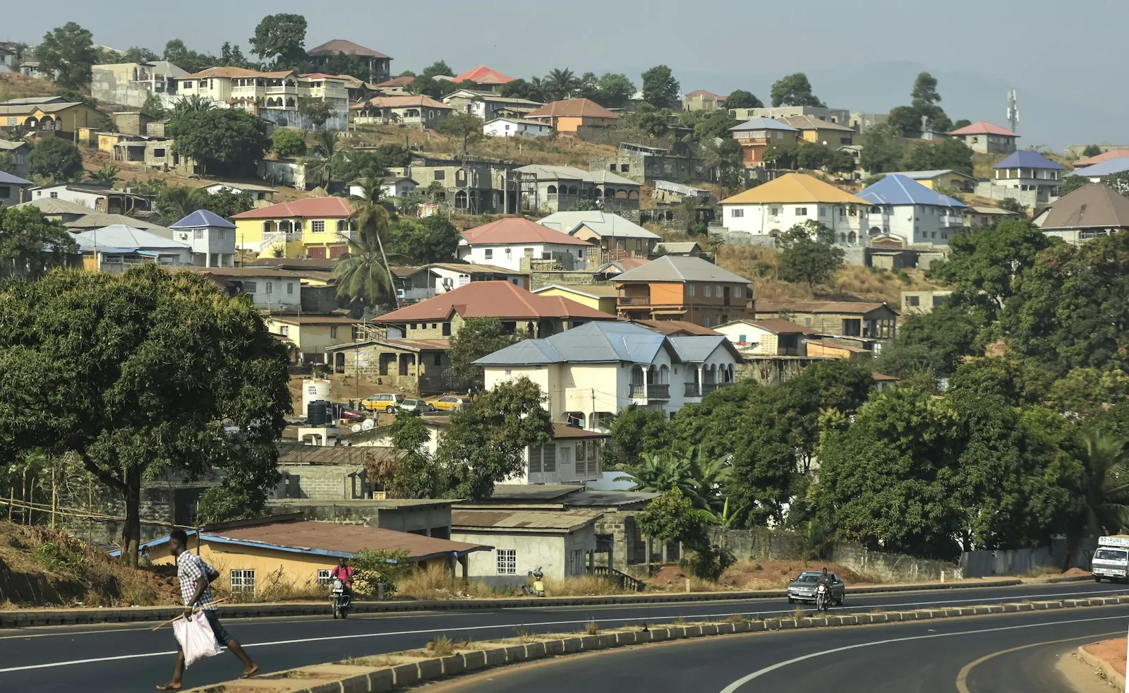 A road curving along a hillside covered in houses and trees in Freetown, with a pedestrian and a car on the road.