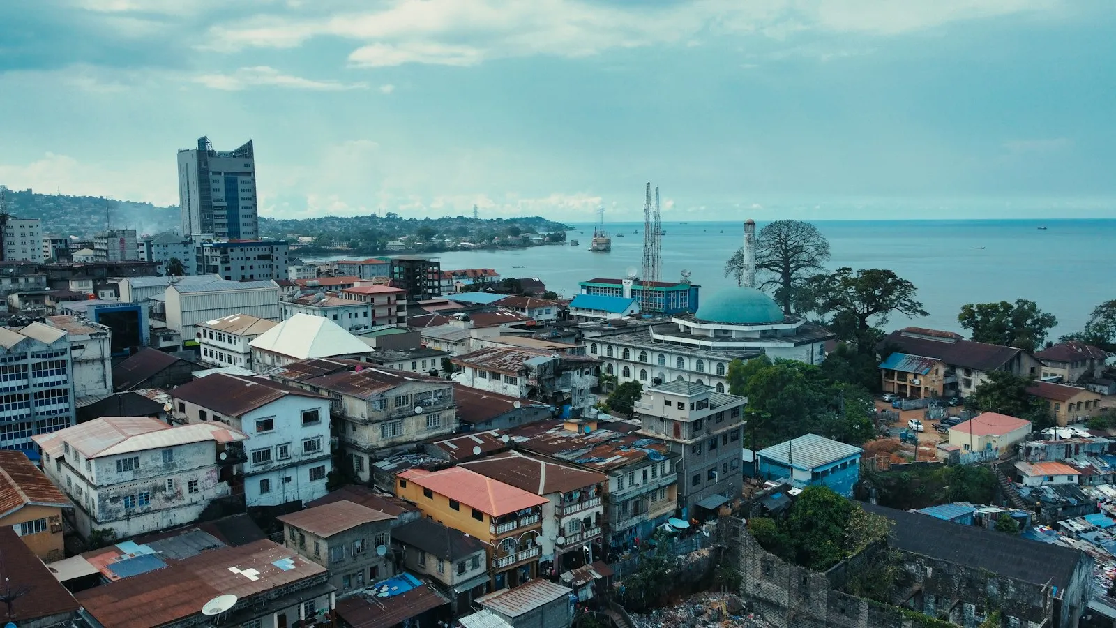 An aerial view of central Freetown, with dense rooftops, a tall office tower and the sea beyond under a blue sky with thin cloud.