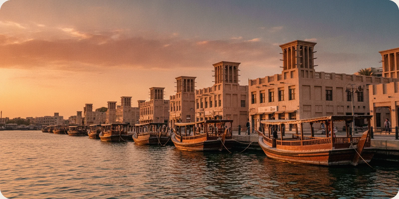 Traditional wooden abra boats moored along Dubai Creek on the Deira side, with low older buildings and wind towers behind in warm late afternoon light.