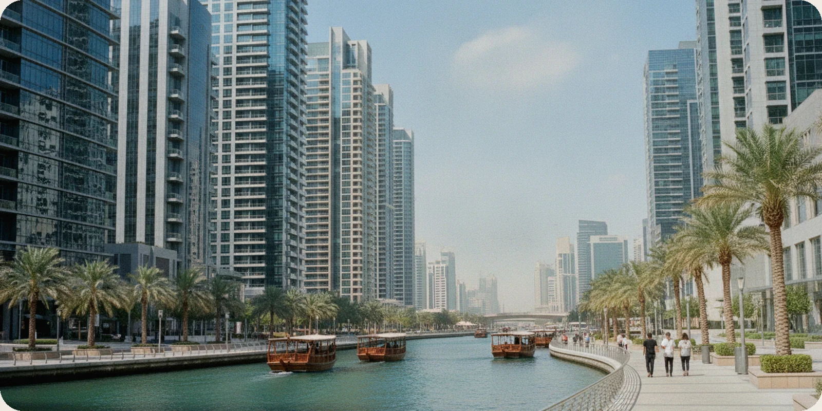 Glass office towers along the Dubai Water Canal in Business Bay, with palm trees, a waterside promenade and small boats under a clear morning sky.