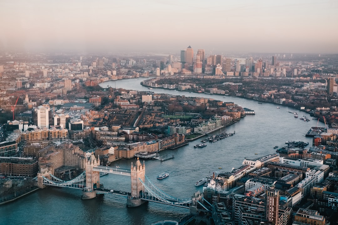 Photographic aerial view of London at golden hour — Tower Bridge in the foreground, the Thames winding through, City of London skyline with the Shard and the Gherkin behind.