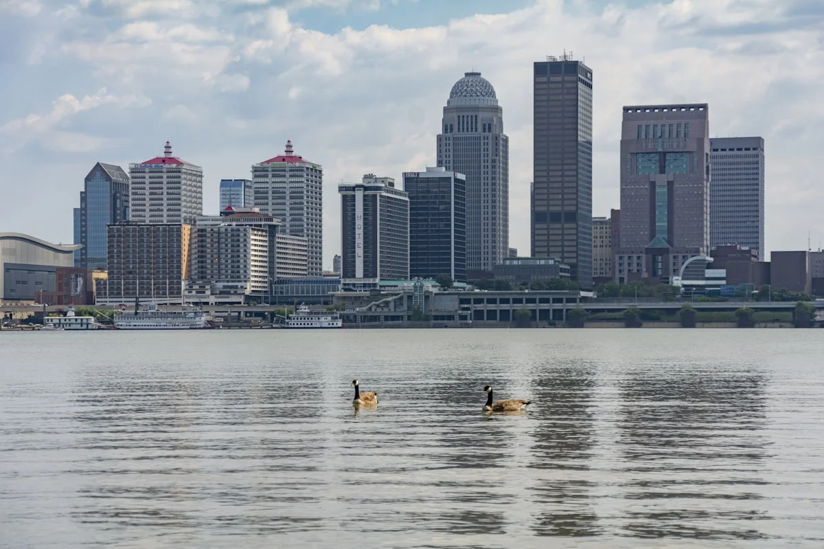 Louisville, Kentucky skyline reflected in the Ohio River at dusk, bridges and downtown office towers visible against a fading sky.
