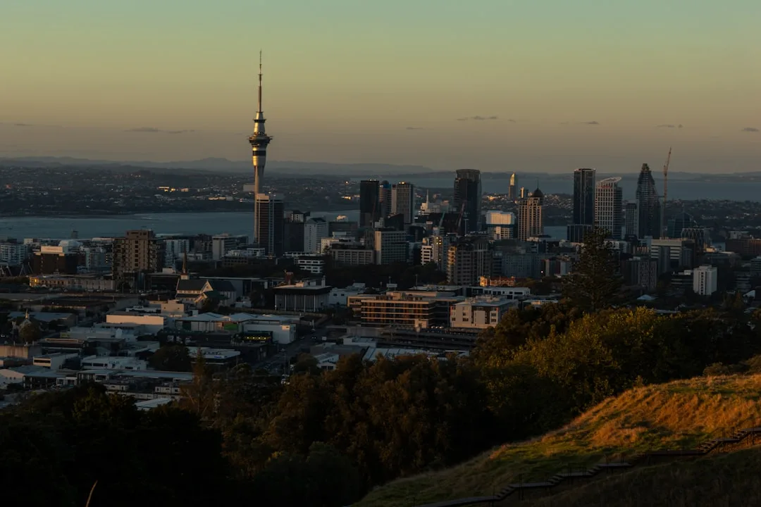 The Auckland waterfront and Sky Tower at golden hour, with sailing boats on a calm harbour below green hills.