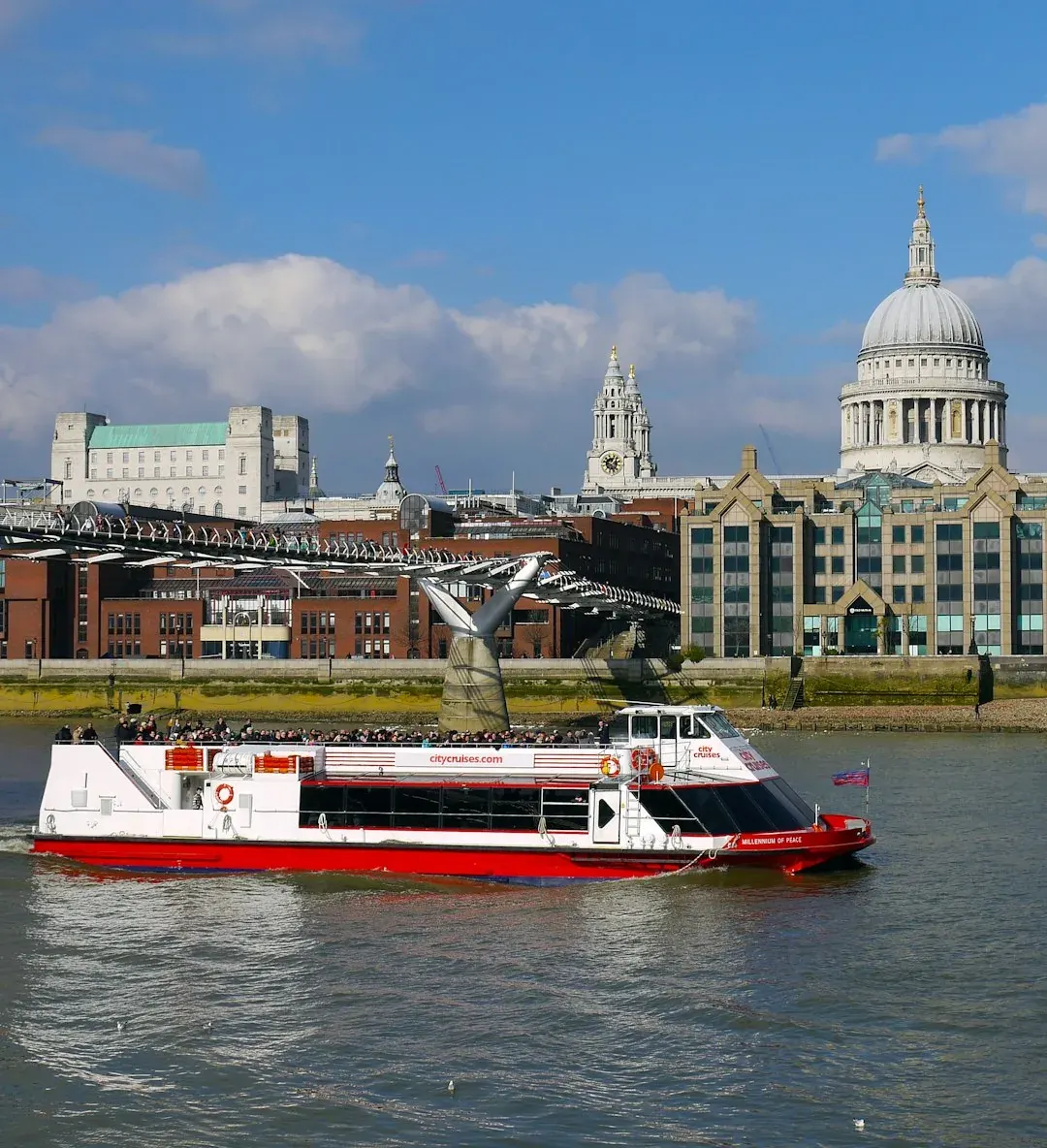 A river cruise boat passing St Paul's Cathedral and the Millennium Bridge on the River Thames in London.